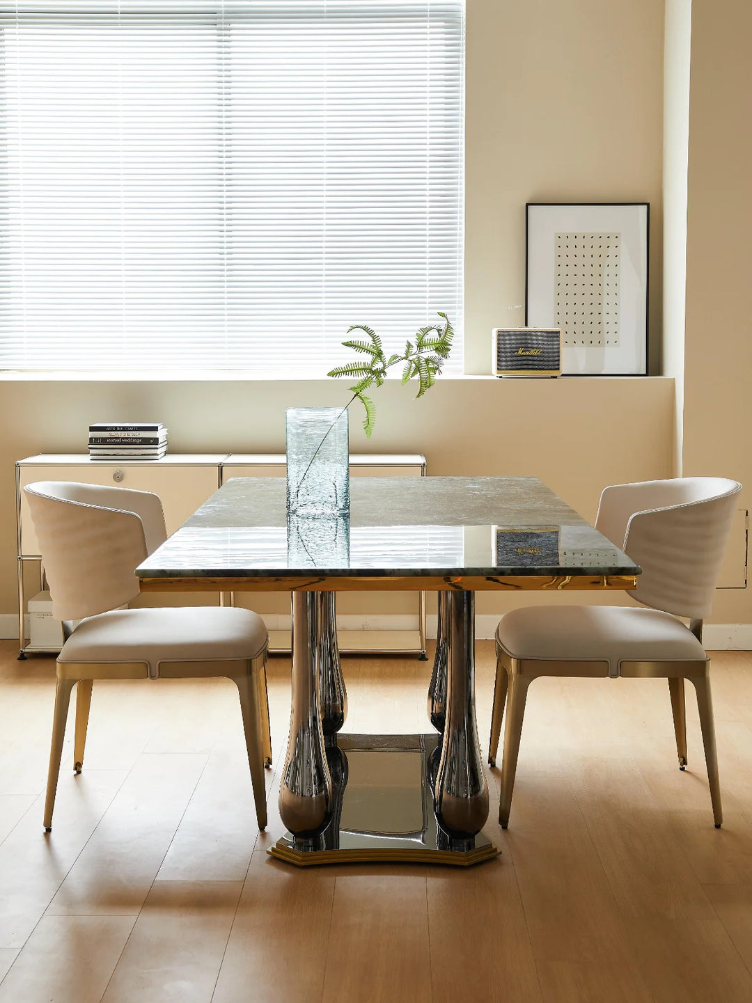 A modern dining table set with a marble top and stainless steel base, accompanied by two chairs with a beige upholstered backrest and seat, set in a room with a light-colored wall and a window.